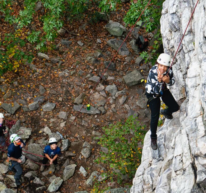 The image shows a group of people rock climbing. A climber is scaling a gray rock face, secured by a rope. Below, two people are on the ground, seemingly belaying or watching the climber. The surrounding area features a forest with fallen leaves and rocks, suggesting an outdoor climbing location. Everyone is wearing helmets for safety.